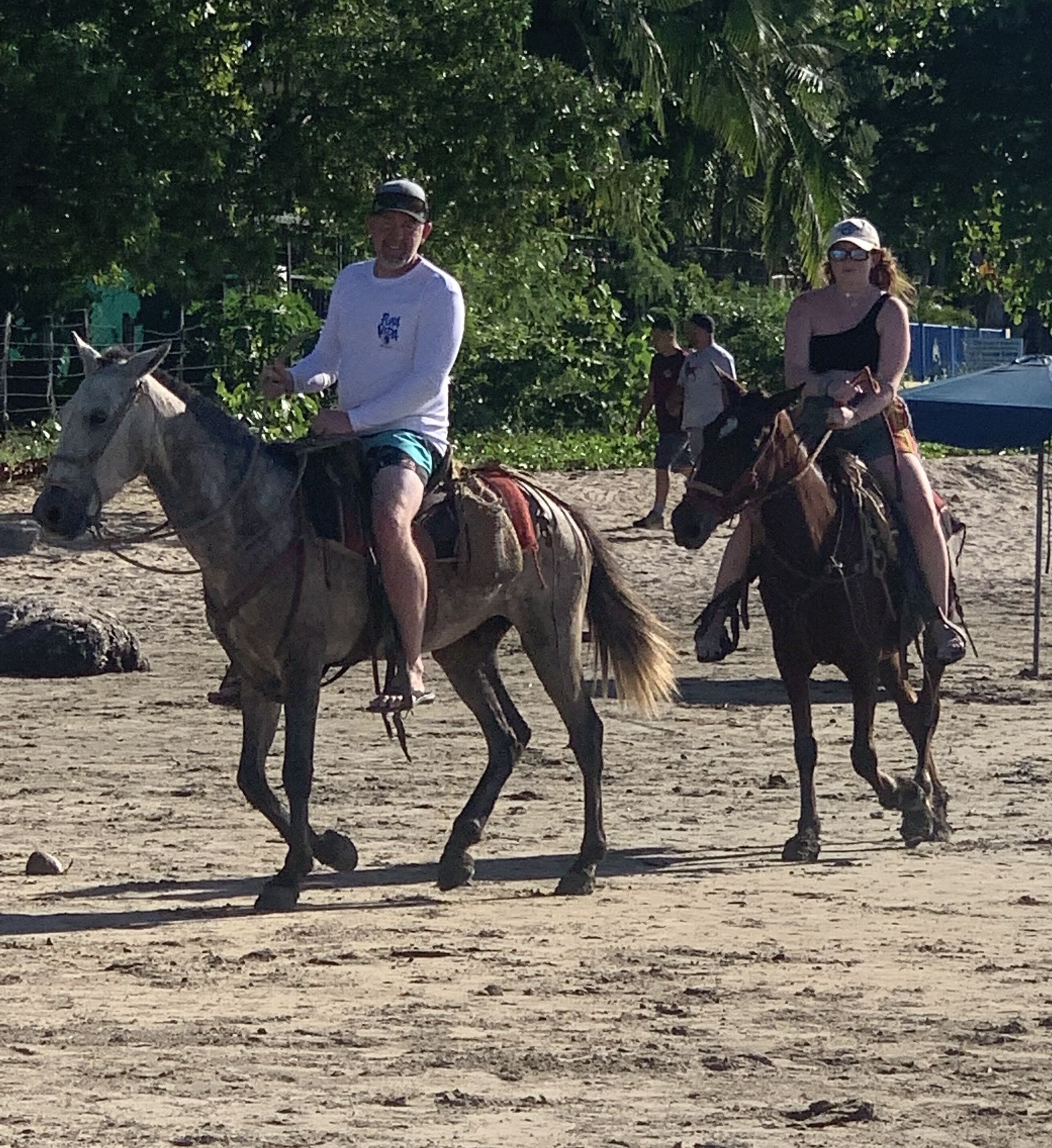 Two people riding horses on a sandy surface with trees in the background.
