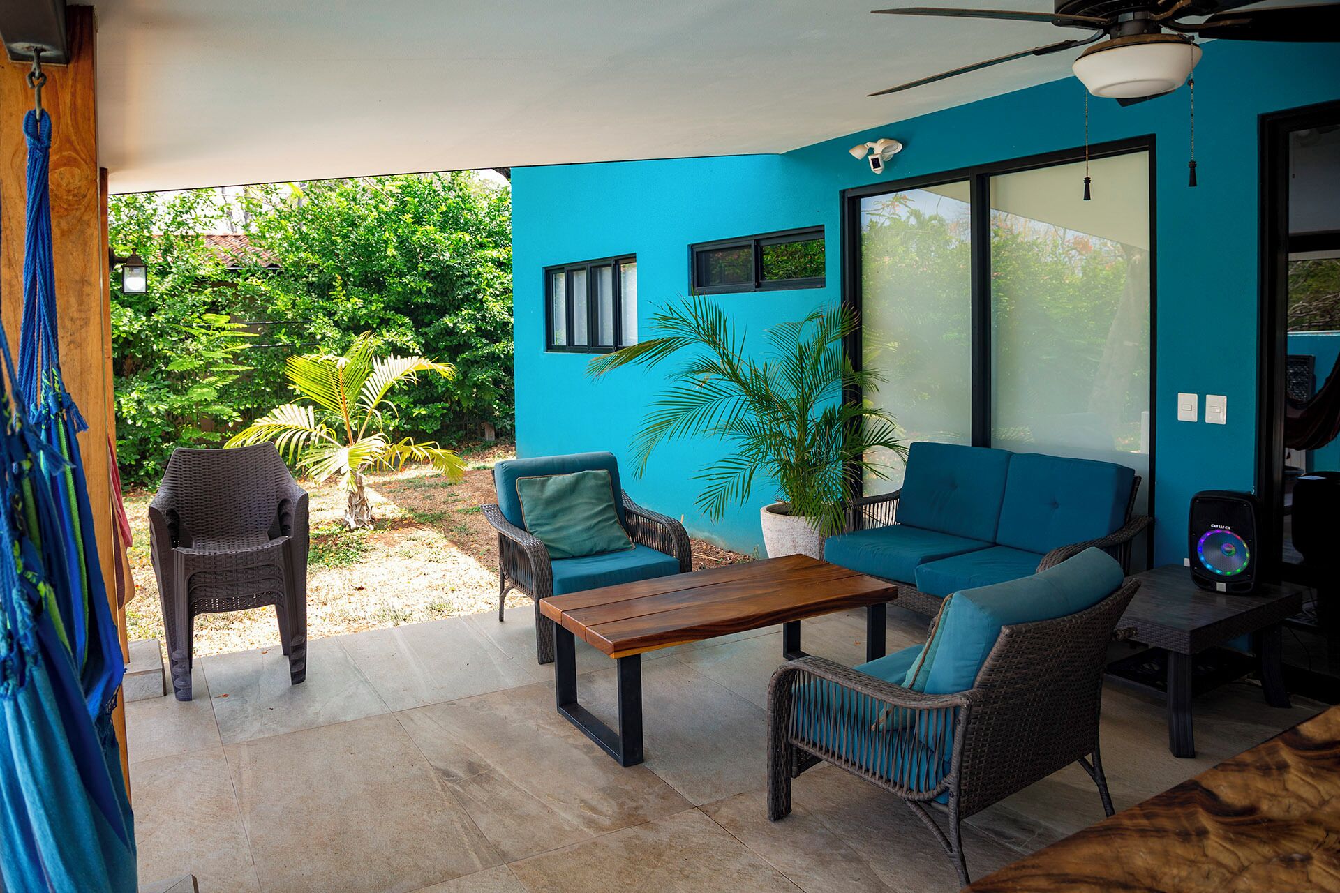Ein überdachter Patio-Bereich mit blauer Wand, Korbmöbeln, einem Holztisch und einem Deckenventilator.