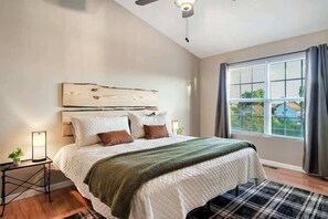 A bedroom with a bed, a headboard, a bedside table with a lamp, a window with curtains, and a ceiling fan.