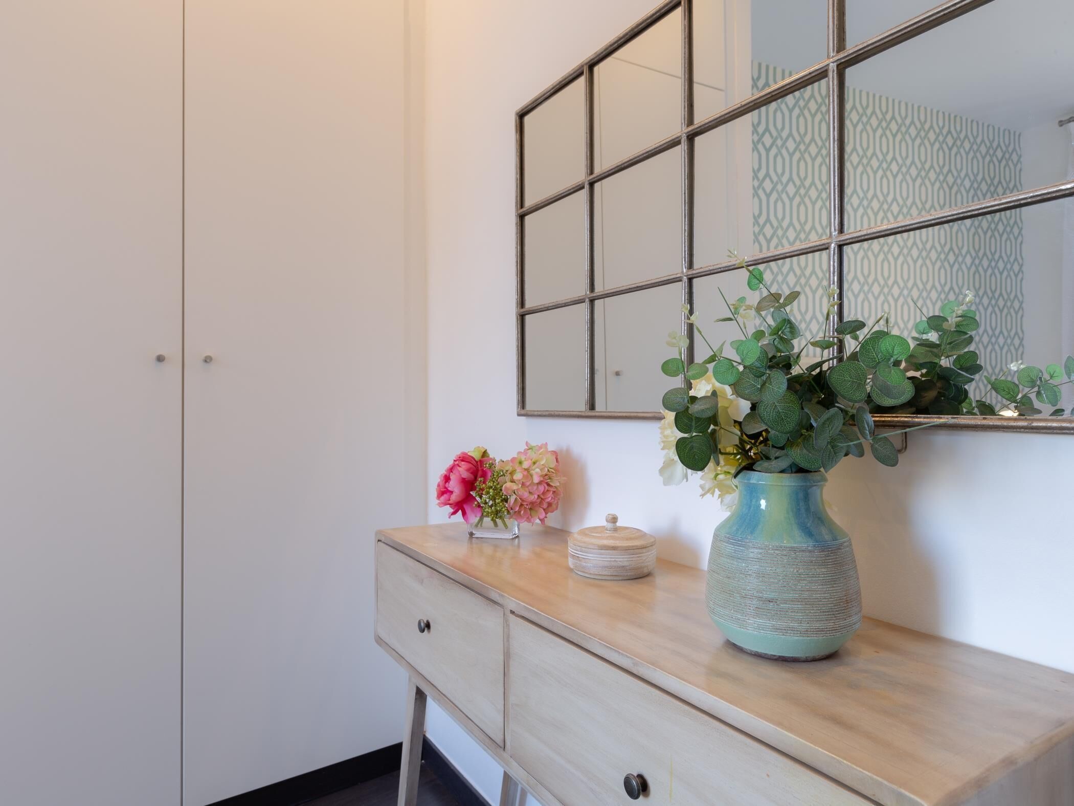 Une salle de bain avec un miroir, une commode agrémentée d’un vase de fleurs et un mur orné d’un motif.
