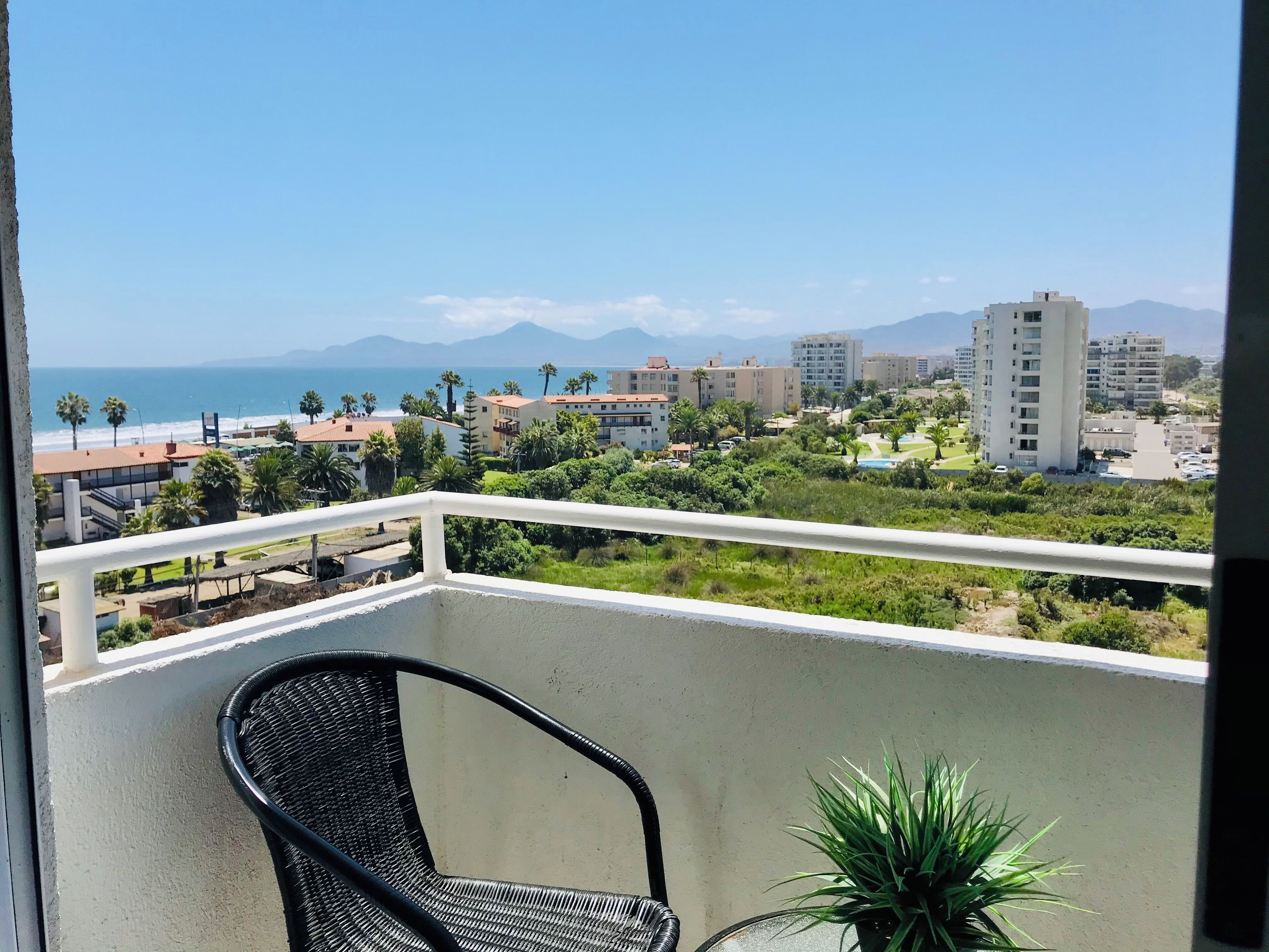 A balcony with a wicker chair and a potted plant, overlooking a coastal cityscape with buildings and mountains in the distance.