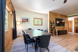 Un vaste espace de vie comprenant une grande table à manger en bois, des chaises sombres, une cheminée et un téléviseur fixé à un mur en bois.