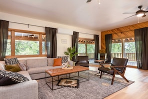 Un salon spacieux comprenant un canapé, des fauteuils, une table basse et une cheminée. De grandes fenêtres offrent une vue sur l’extérieur.