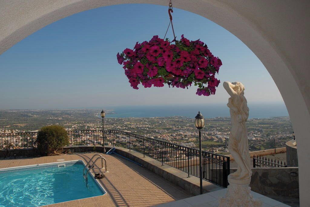 A pool area with a view of a city and the sea, a hanging flower basket, and a statue.