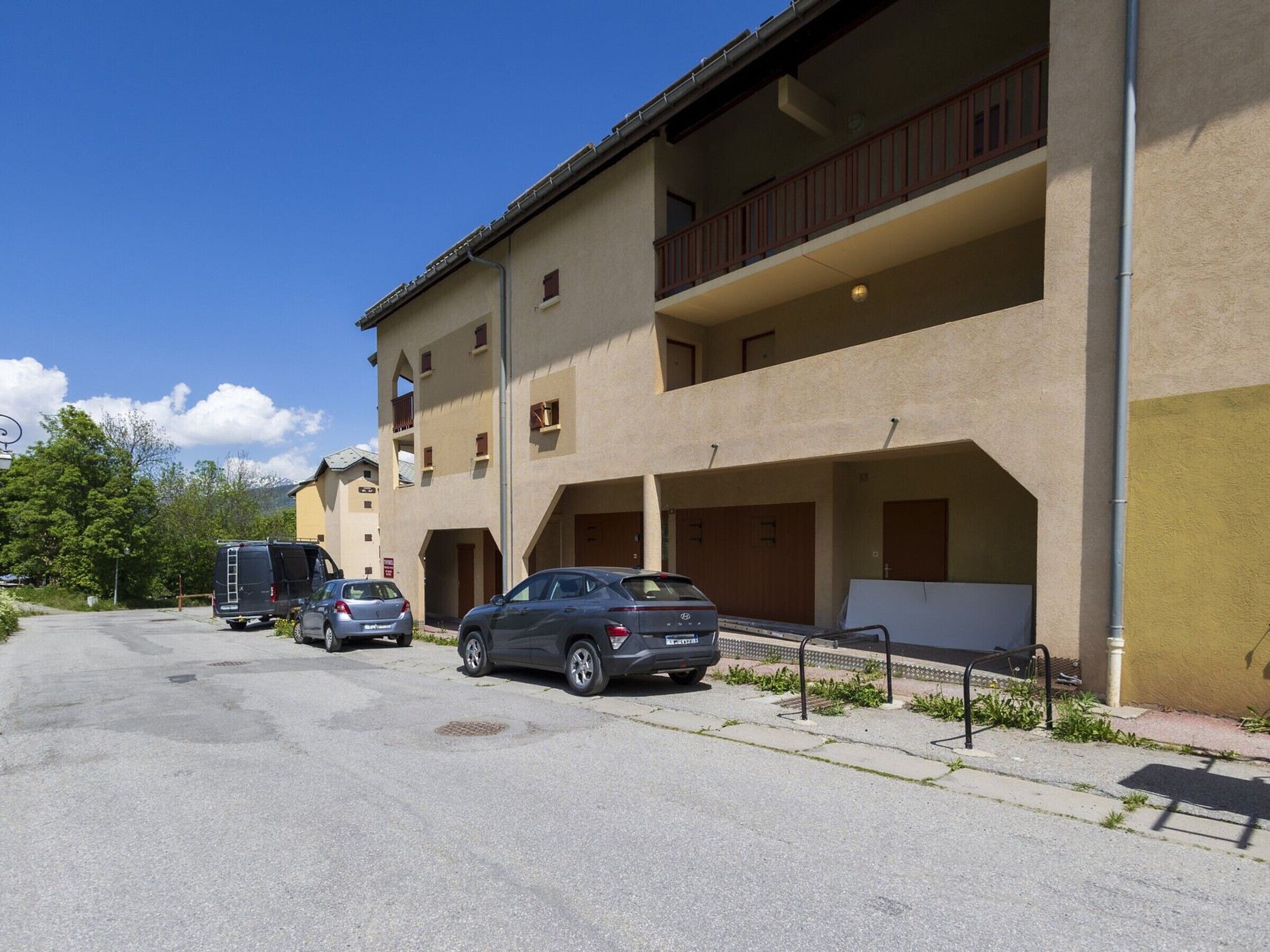 A multi-story building with balconies, a parking area with cars, and a clear sky.