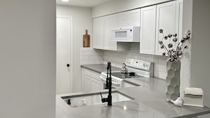 A modern kitchen with white cabinetry, a central island with a sink, two black bar stools, a microwave, and a vase with dried flowers. - NEW REMODEL in the Heart of Eagle near BOISE (Eagle)