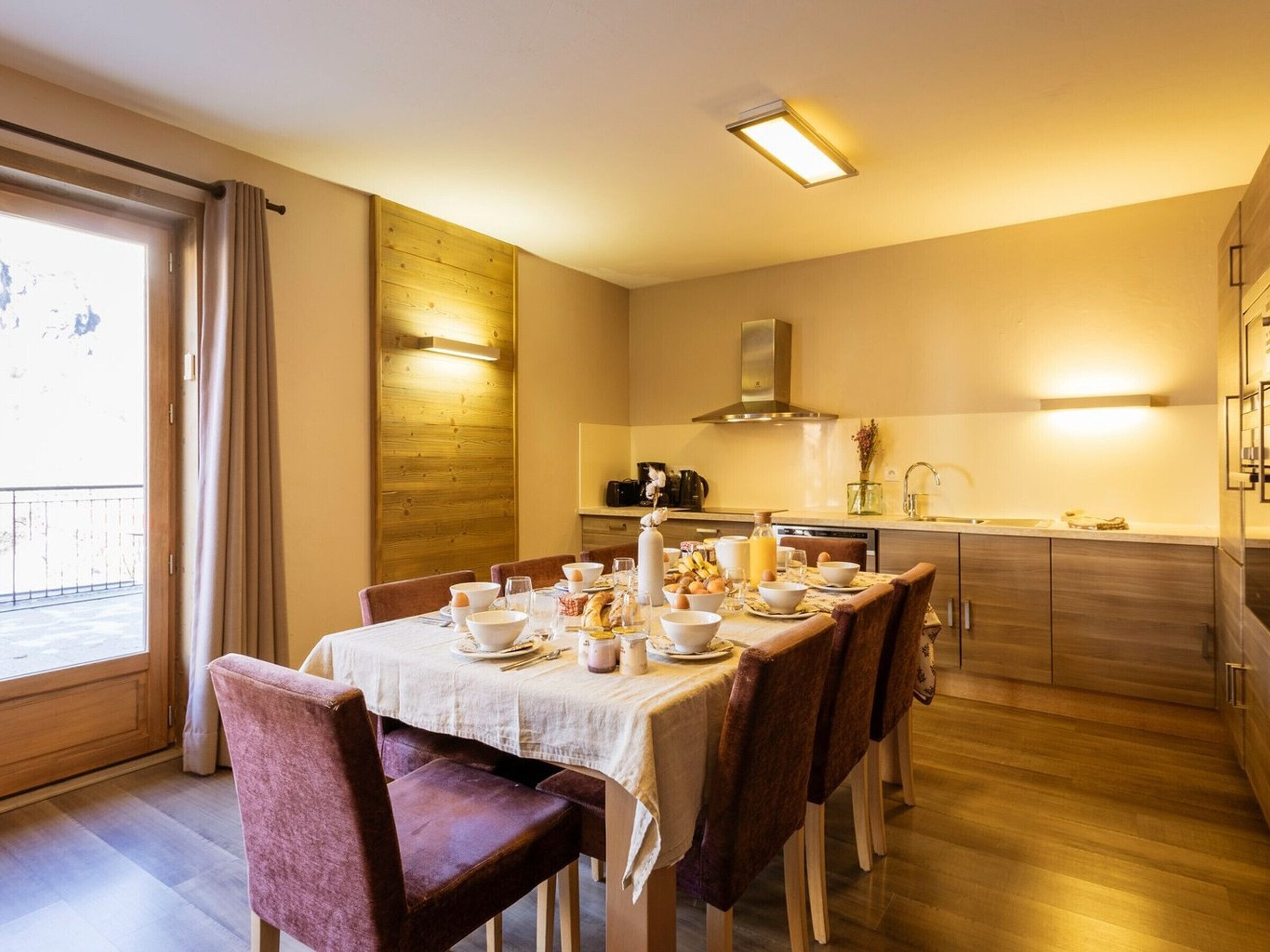 Zona de dinar amb taula preparada per l'esmorzar, sòl de fusta, cuina al fons i vista per una finestra.