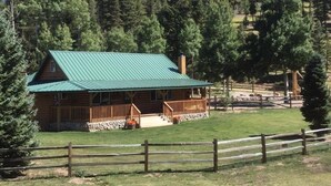 A log cabin with a green roof, surrounded by a wooden fence and a grassy yard.