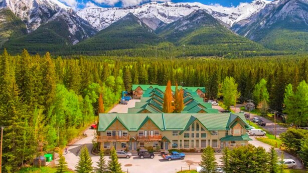 A lodge with a green roof, surrounded by dense forest and snow-capped mountains in the background.