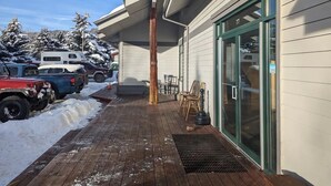 En träaltan med snötäckt mark, en byggnadsingång med en glasdörr och parkerade fordon.