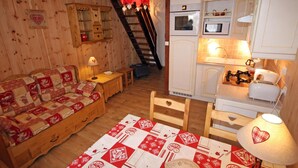 A cozy room with a wooden floor, a quilted sofa, a table with a patterned tablecloth, a lamp, a kitchen area with a stove, oven, and microwave, and a staircase leading to an upper level. - Duplex 3 rooms 4 people, south-facing terrace, private parking (Valloire)