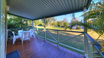 A covered outdoor area with a wooden deck, white plastic chairs, and a view of a grassy area with trees.