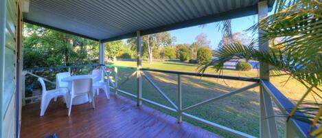 A covered outdoor area with a wooden deck, white plastic chairs, and a view of a grassy area with trees.