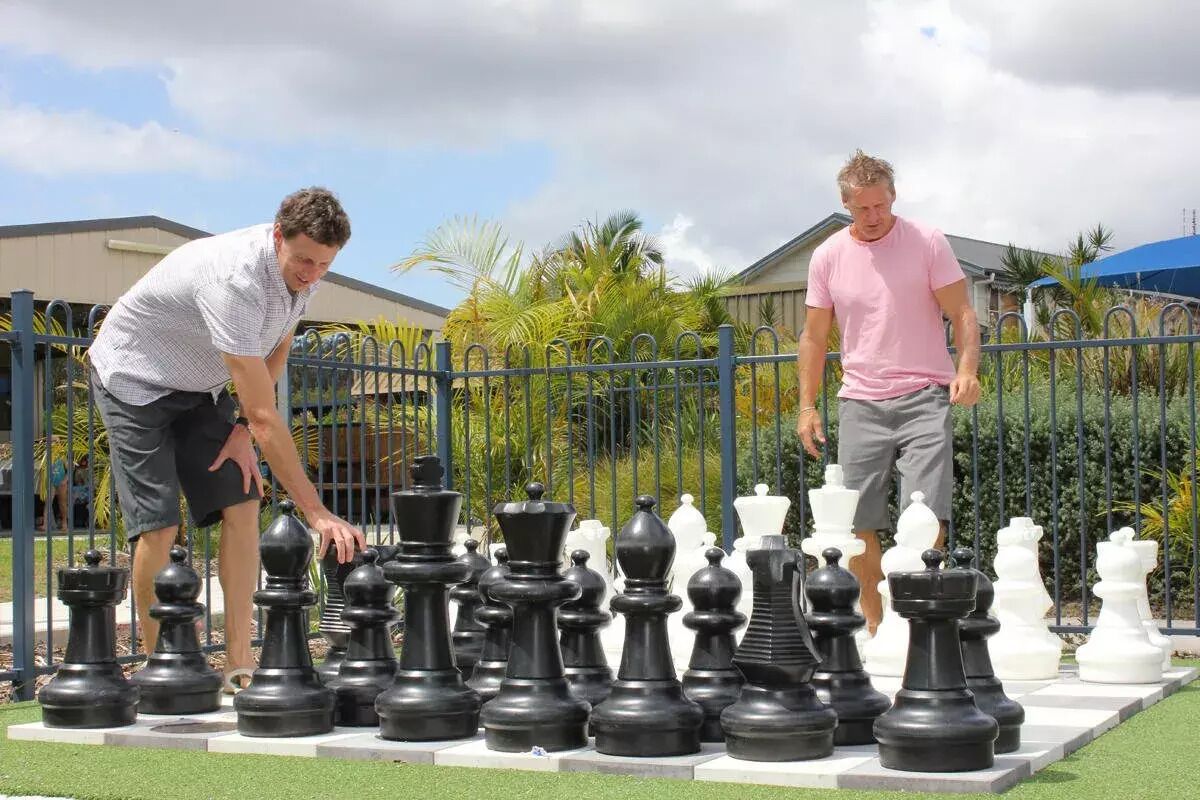 Two individuals playing chess on a large outdoor board surrounded by a metal fence.