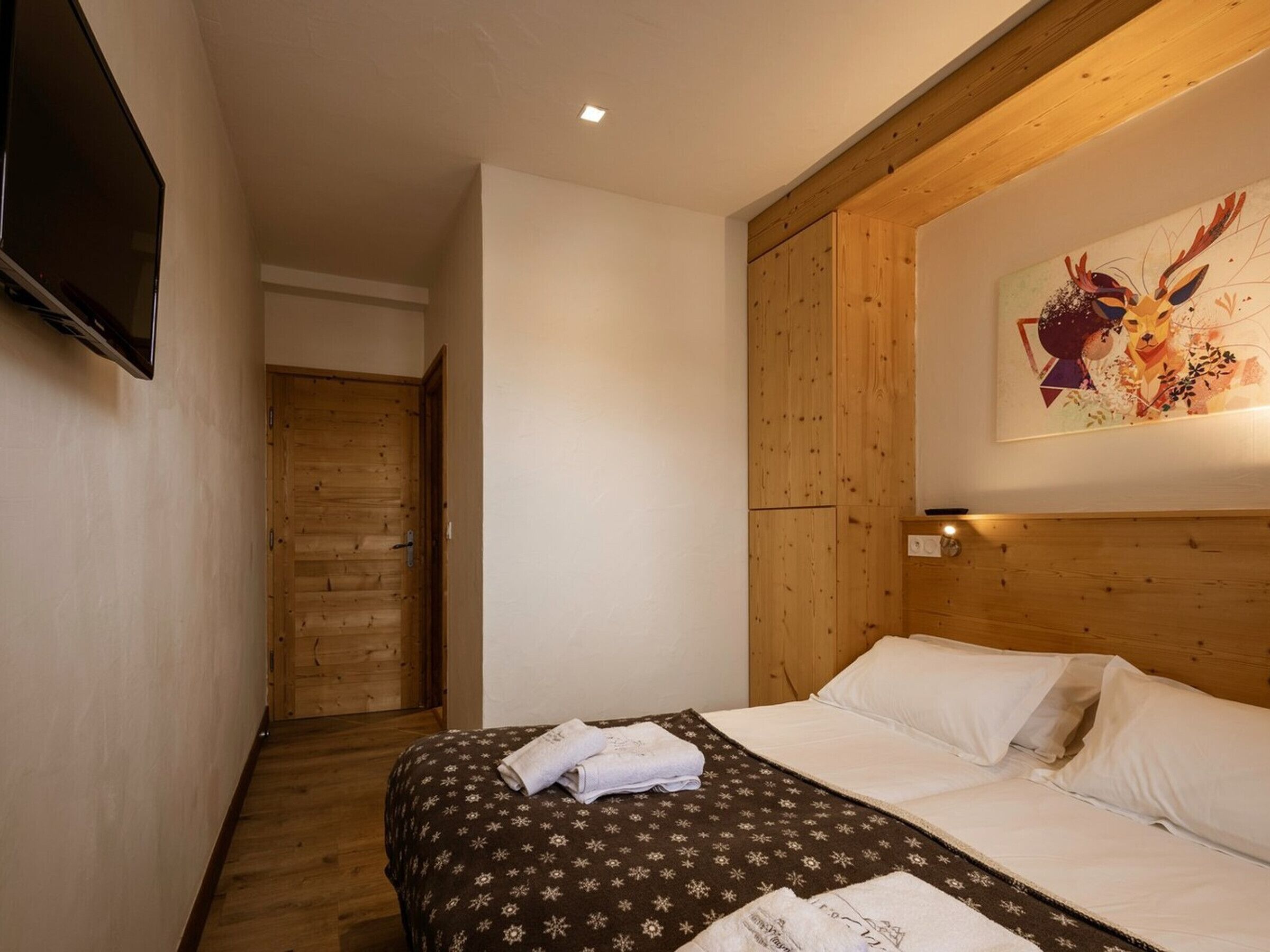 A hotel room with a bed, a wooden door, a painting on the wall, and a television mounted on the wall.