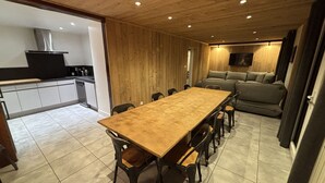 En rummelig spiseplads med et langt træbord og stole, et køkken med moderne hårde hvidevarer, og en hyggelig opholdsstue med sofa og TV.