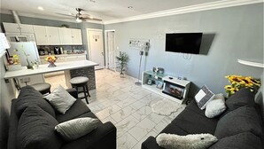 Una sala de estar moderna con un sofá gris, un televisor de pantalla plana en la pared, una cocina con armarios blancos y un comedor con un taburete de bar.