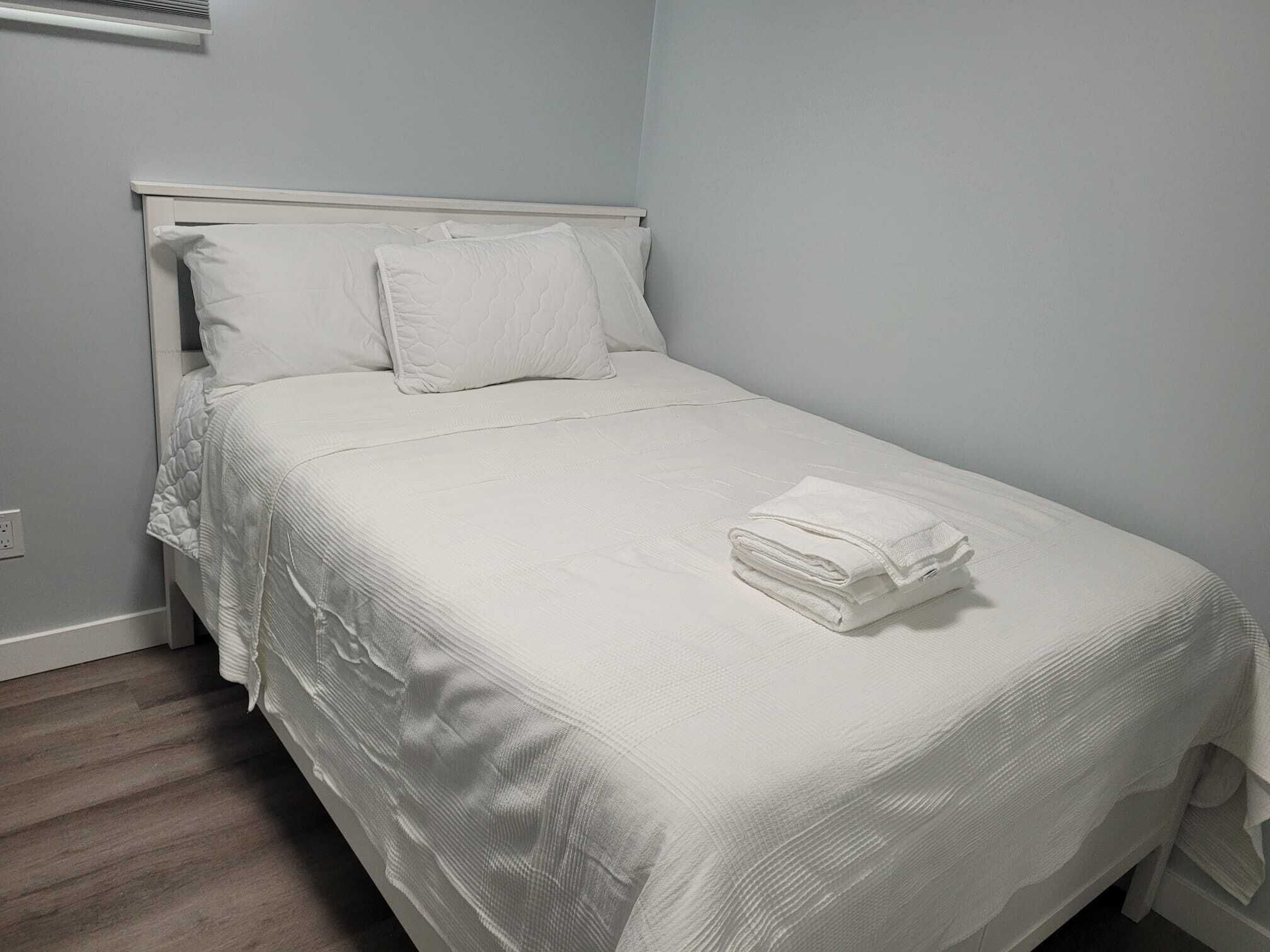 A neatly made bed with a white bedspread and pillows, a folded white towel on the bed, and a white headboard.