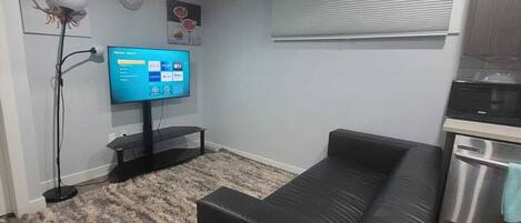 A modern living room with a black sofa, a flat-screen TV, a standing lamp, and a kitchen area with a stainless steel appliance.
