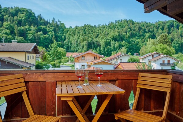 A wooden deck with a table and chairs, overlooking a forested hillside with houses. - Spacious 72 m² vacation apartment, sleeps 1–4, 2 bedrooms, Wi-Fi, balcony (Marquartstein)