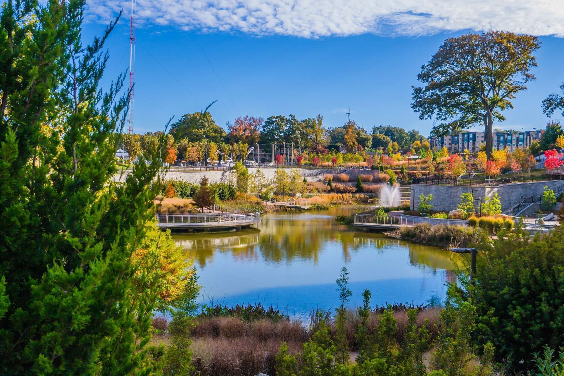 A serene lakeside park with a bridge, trees, and a building in the background.