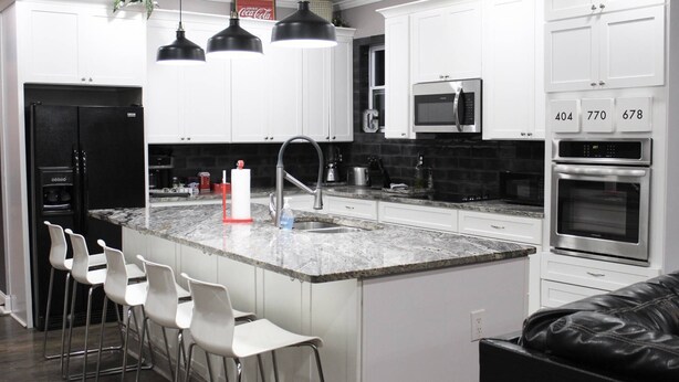 Una cocina moderna con armarios blancos, un refrigerador negro, un microondas integrado y un horno doble. Hay tres taburetes blancos y un sofá de cuero negro.