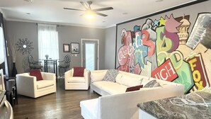 Una sala de estar con un sofá blanco, una mesa de comedor con sillas y un mural grande en la pared.