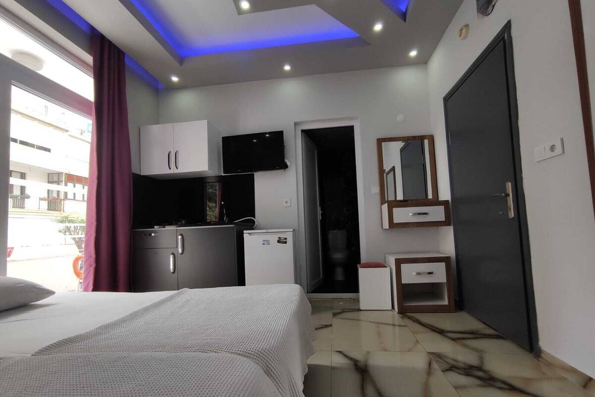 A modern hotel room with a bed, a small kitchenette, a mirror, and a door.