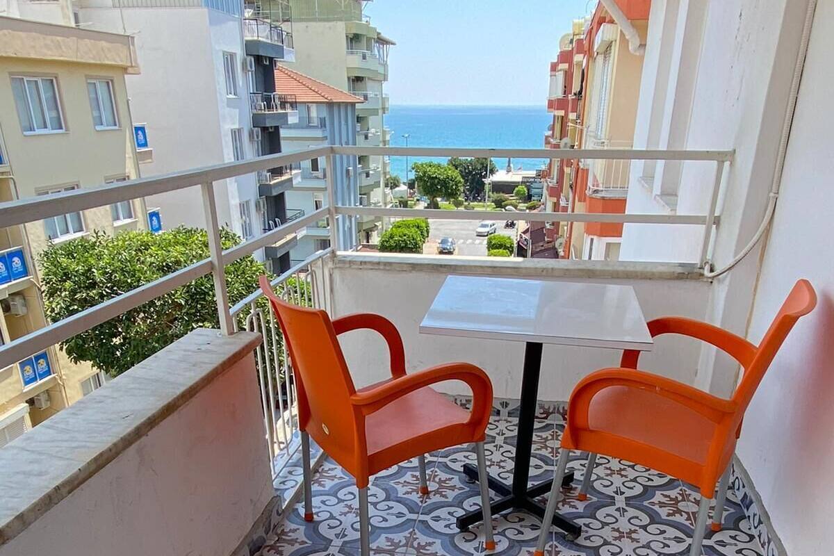 A balcony with two orange chairs and a table, patterned floor tiles, and a view of the sea and buildings.