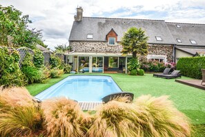 Una casa de piedra con piscina, un porche con paredes de vidrio y un césped bien cuidado.
