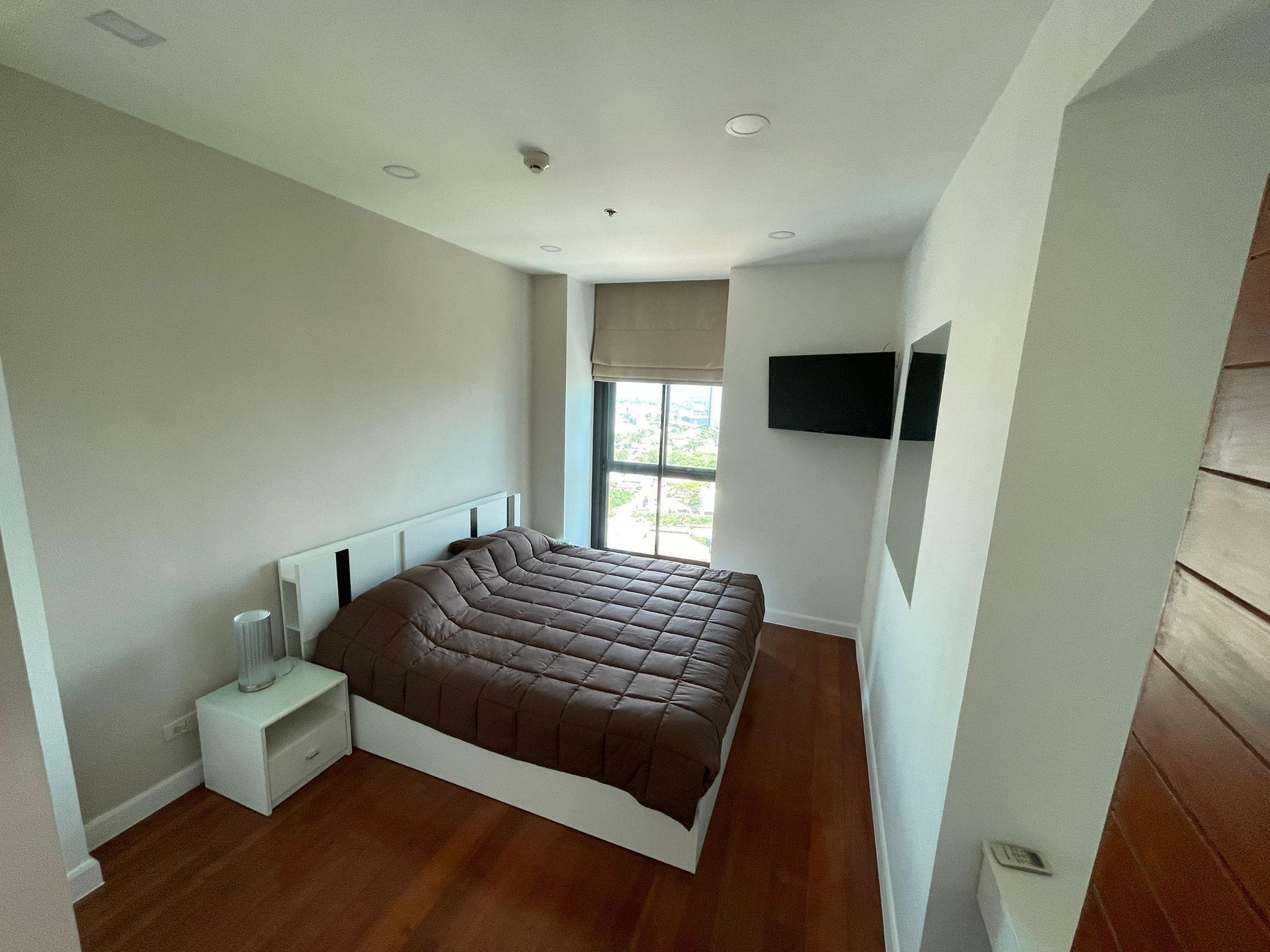 Une chambre avec un grand lit, une table de chevet, un téléviseur fixé au mur et une fenêtre donnant sur un paysage verdoyant.