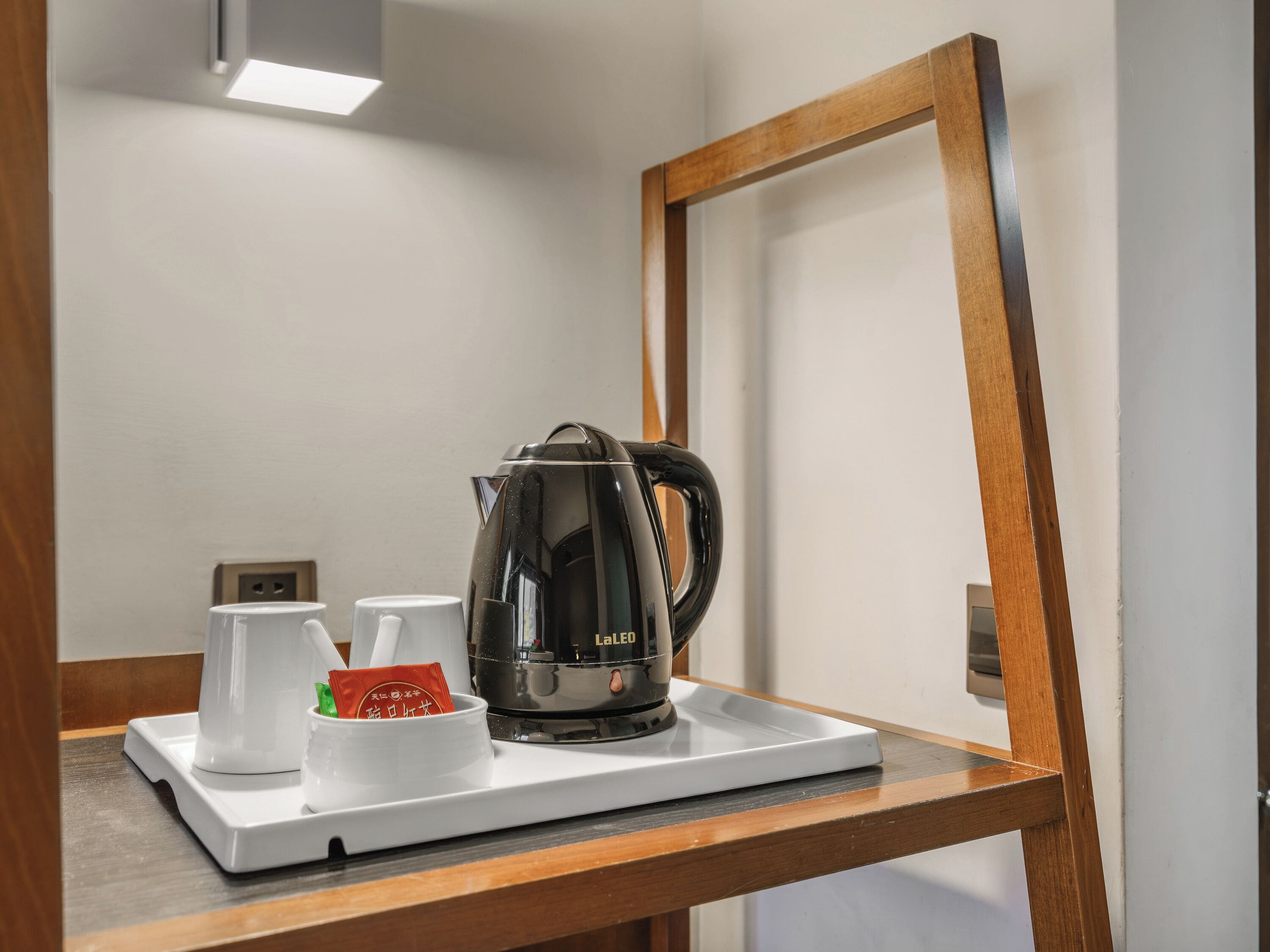 A LaLeu coffee maker on a white tray with two white cups and a red packet, placed on a wooden table in a room with a white wall and a door frame.