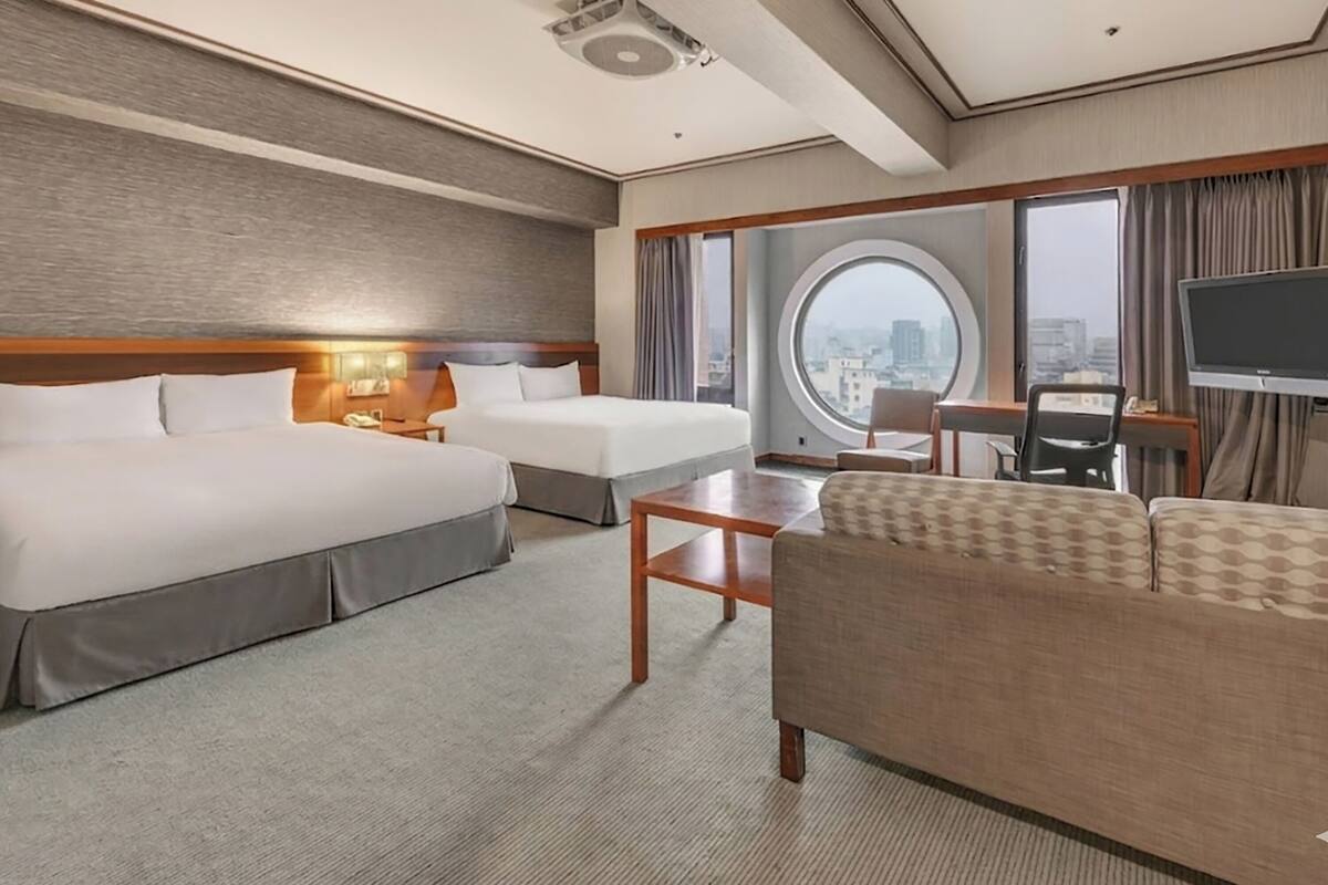 A hotel room with two beds, a sofa, a desk, a TV, and a large window with a city view.