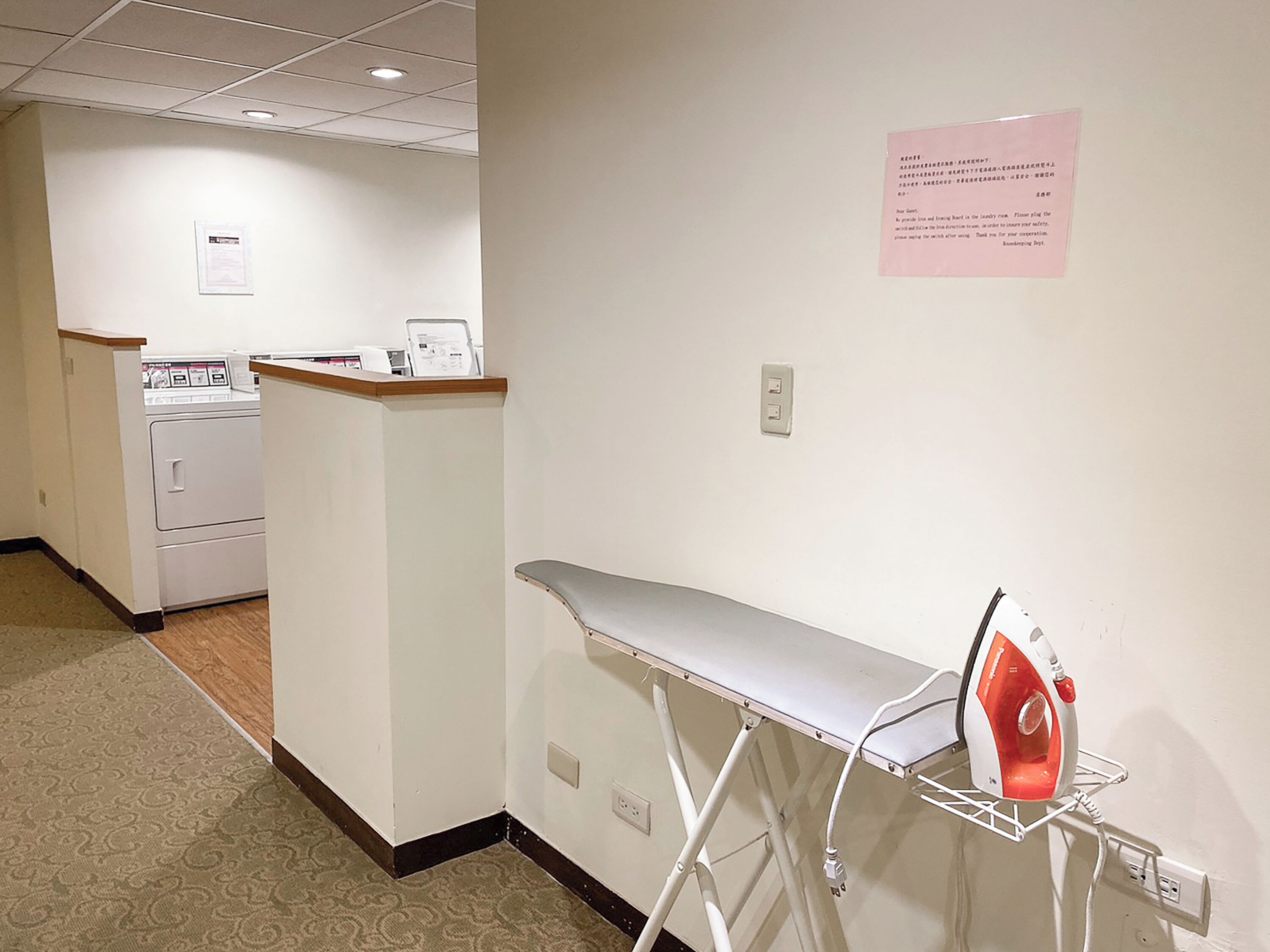 A hotel room with a washing machine, ironing board, and a notice on the wall.