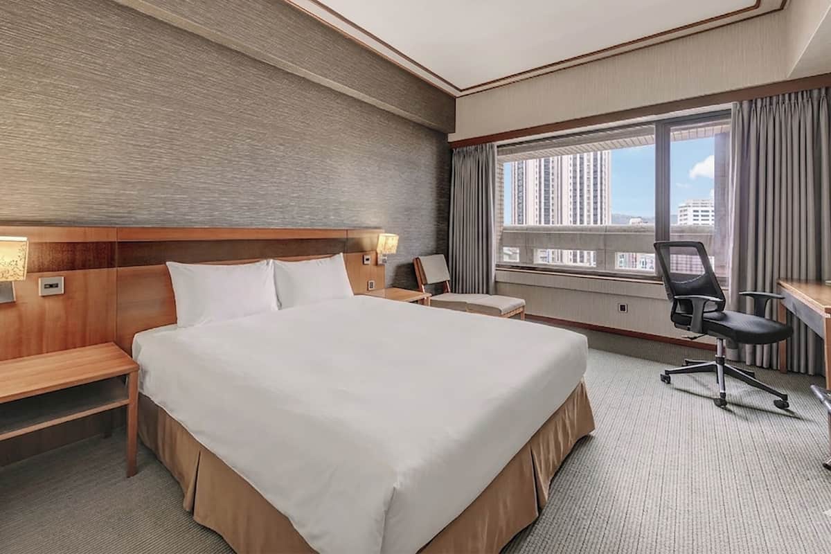 A hotel room with a large bed, a desk, a chair, and a window with a city view.