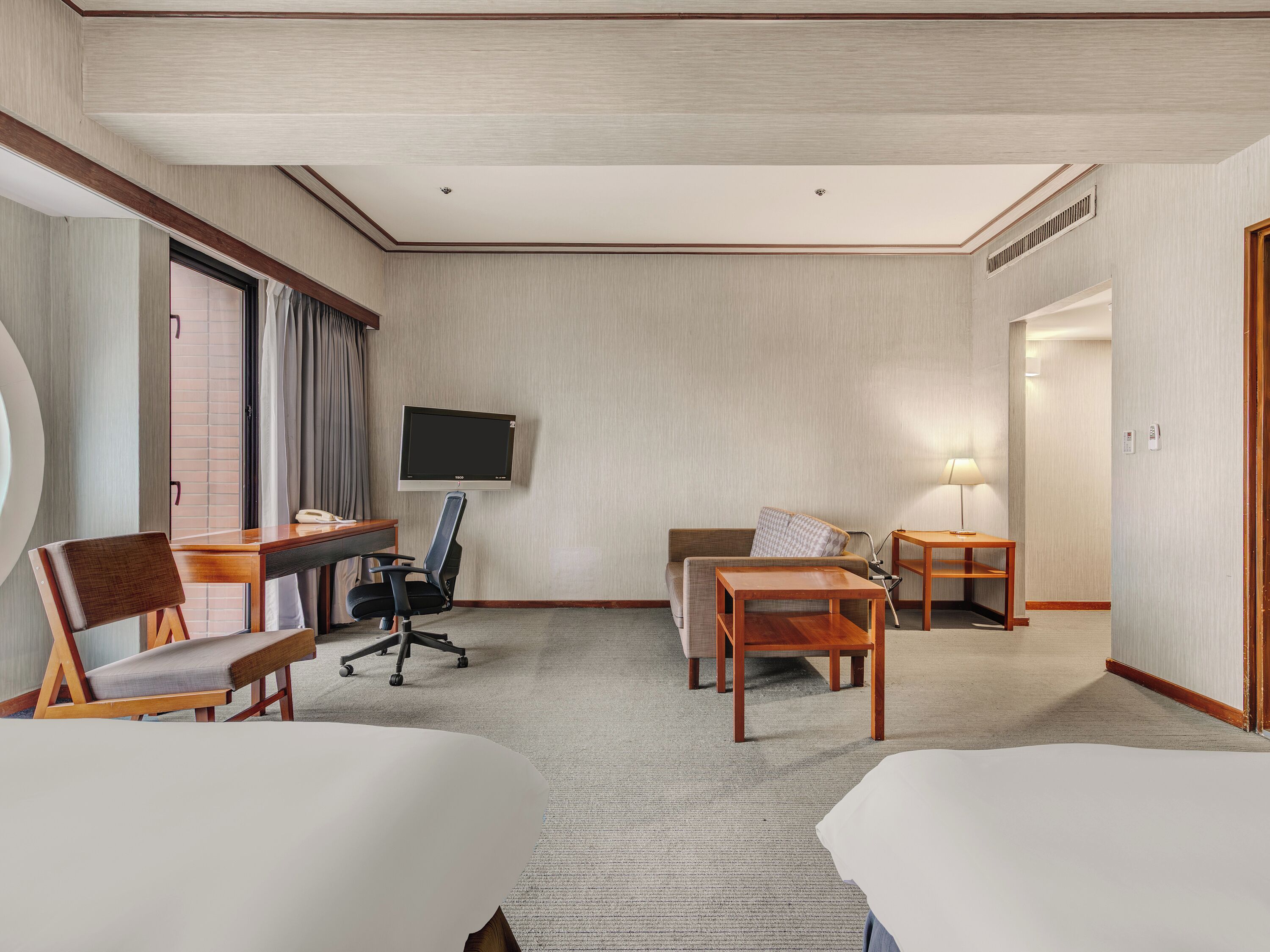 A hotel room with a flat-screen TV, two beds, a desk with a chair, a sofa, and a small table with a lamp.