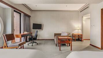 A hotel room with a flat-screen TV, two beds, a desk with a chair, a sofa, and a small table with a lamp.
