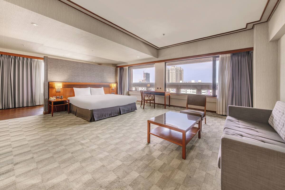 A hotel room with a bed, a desk, a sofa, and a view of the cityscape.