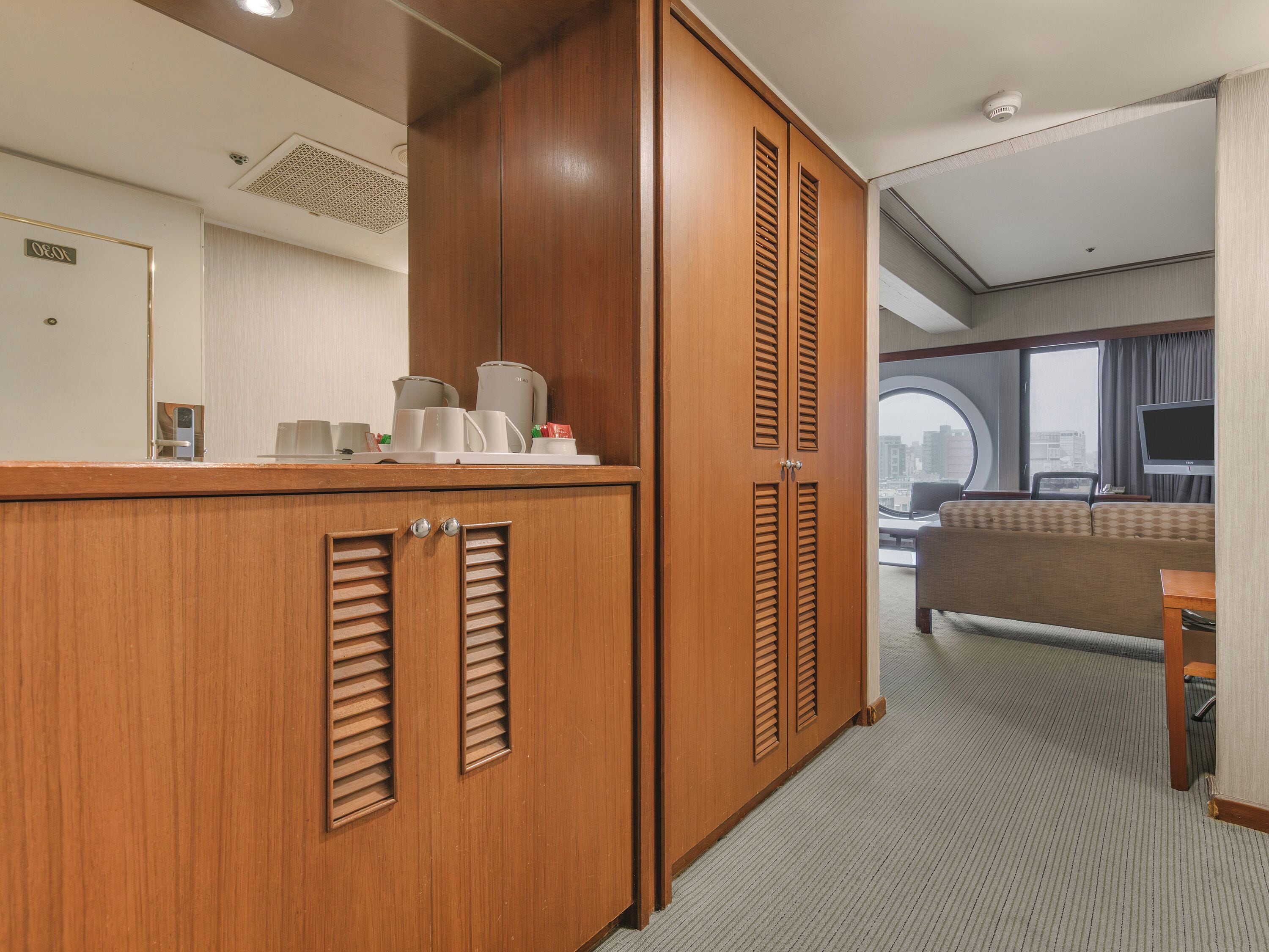 A hotel room with a wooden wardrobe, a kitchenette, a sofa, and a television.