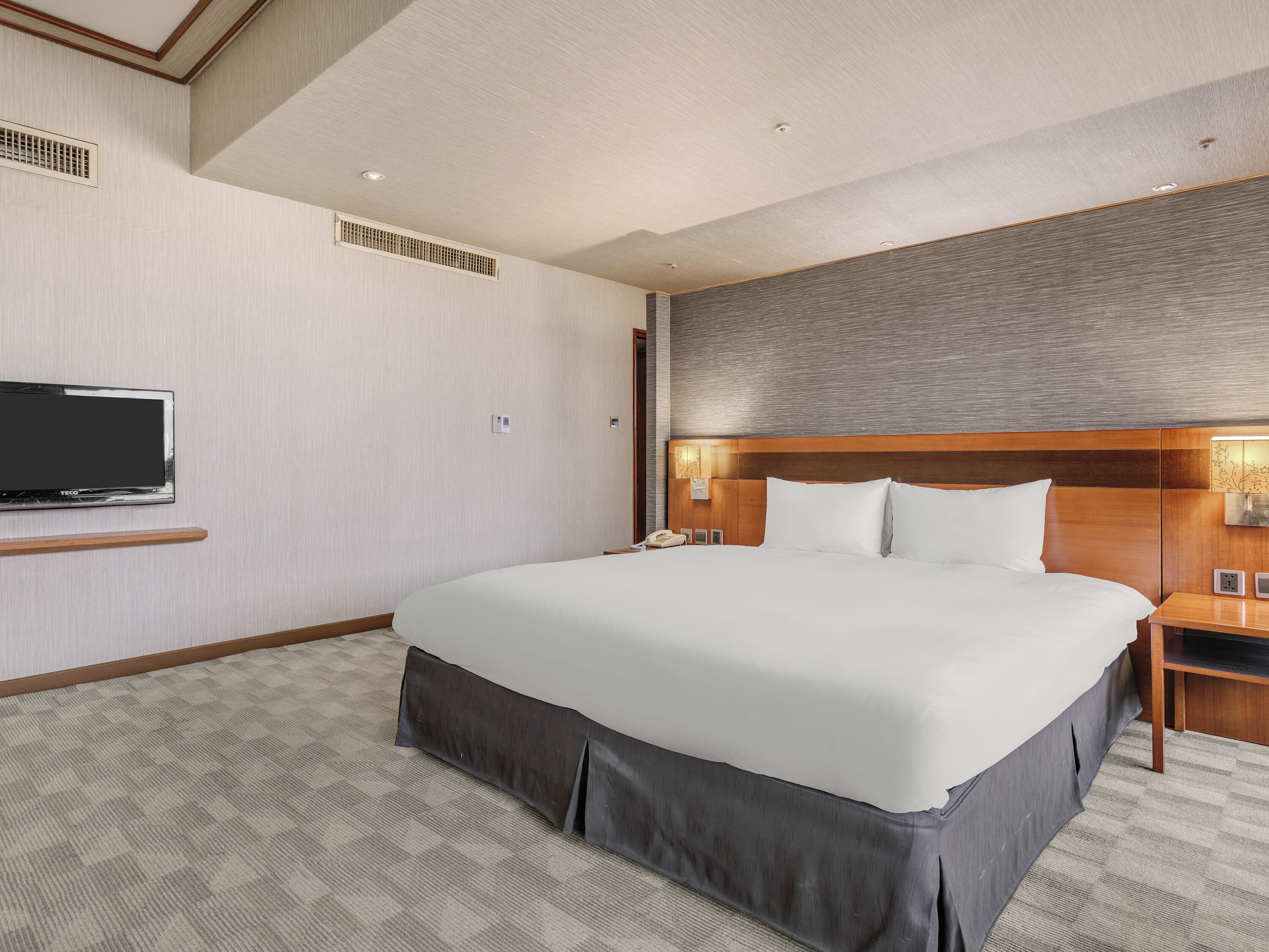 A hotel room with a large bed, a TV mounted on the wall, a bedside table, and a headboard with built-in lighting.