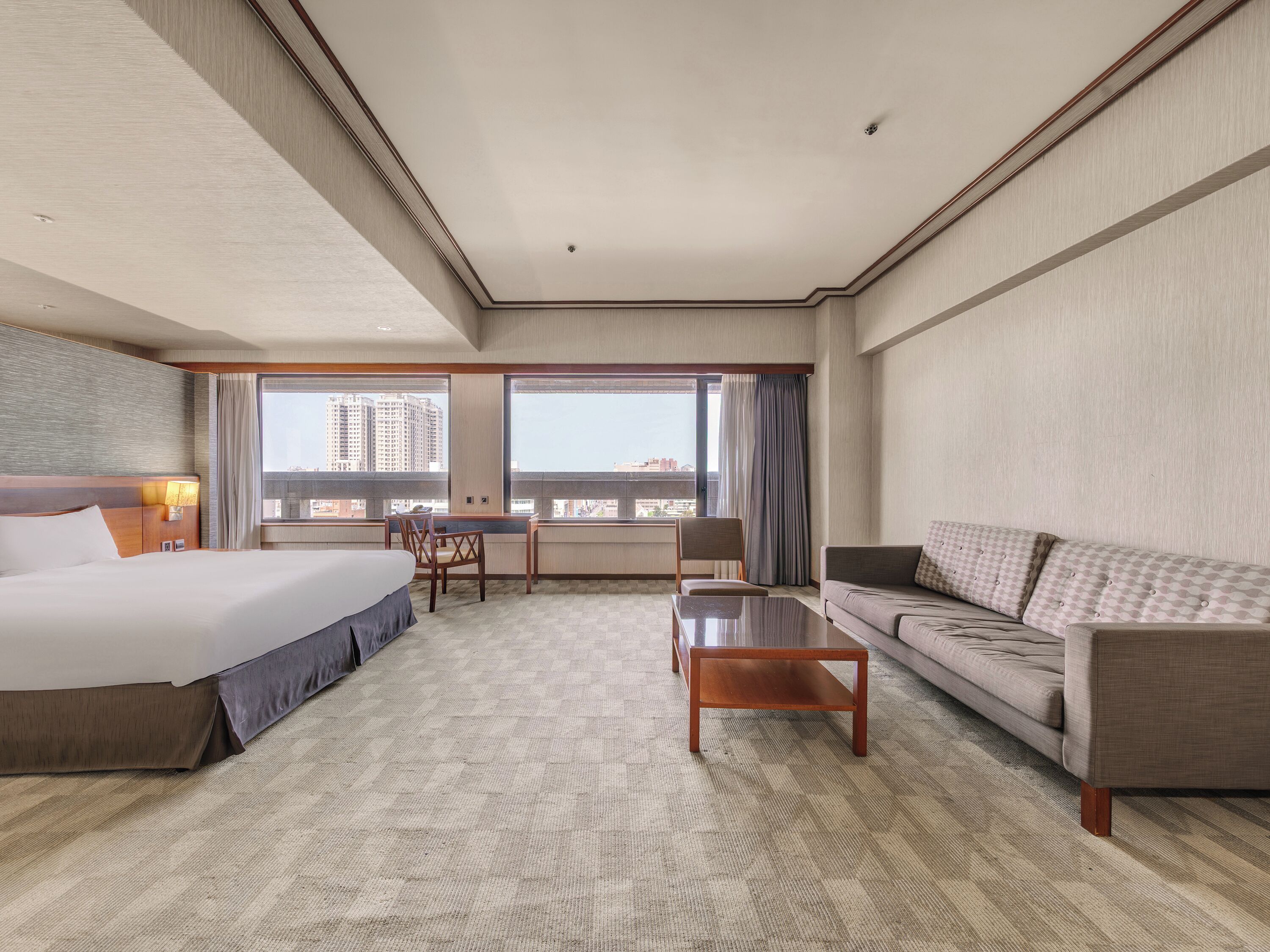 A hotel room with a bed, a sofa, a small table, and a view of the city through large windows.