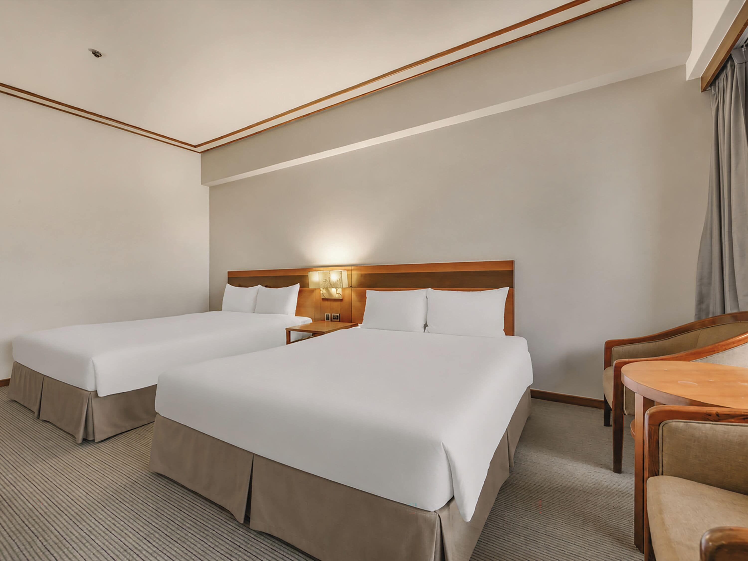 A hotel room with two beds, a wooden headboard, a bedside table, a chair, and a lamp.