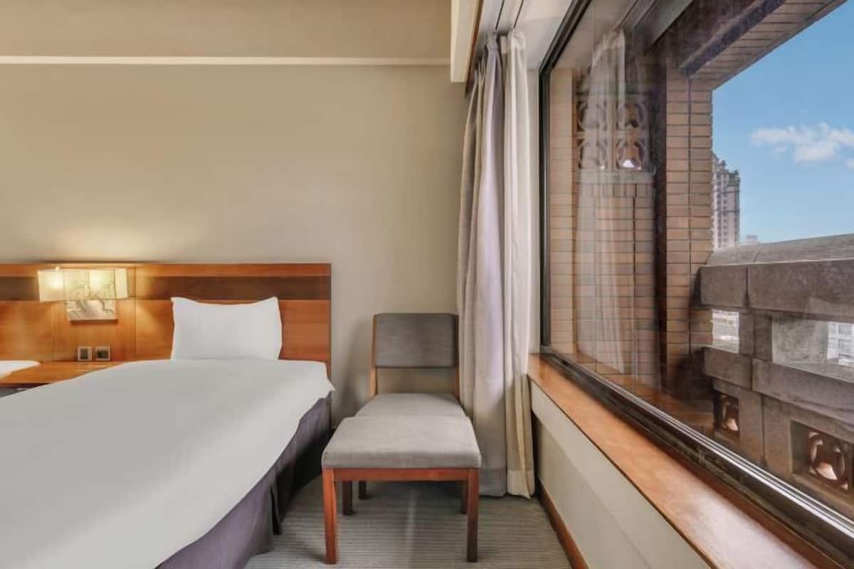 A hotel room with a bed, a chair, a window with a view of buildings, and a wall-mounted lamp.