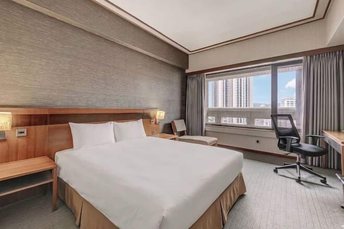 A hotel room with a large bed, a desk, a chair, a bench, and a window with a view of buildings.