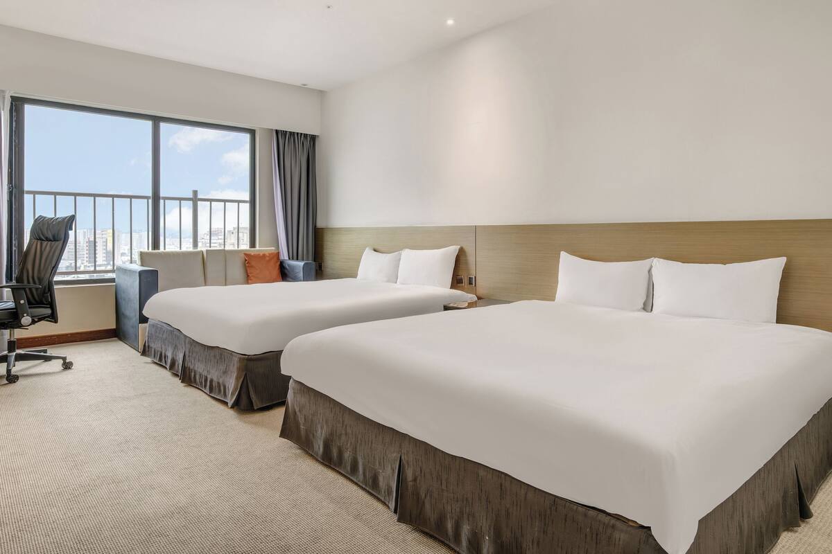A hotel room with two beds, a desk, a chair, and a large window with a city view.