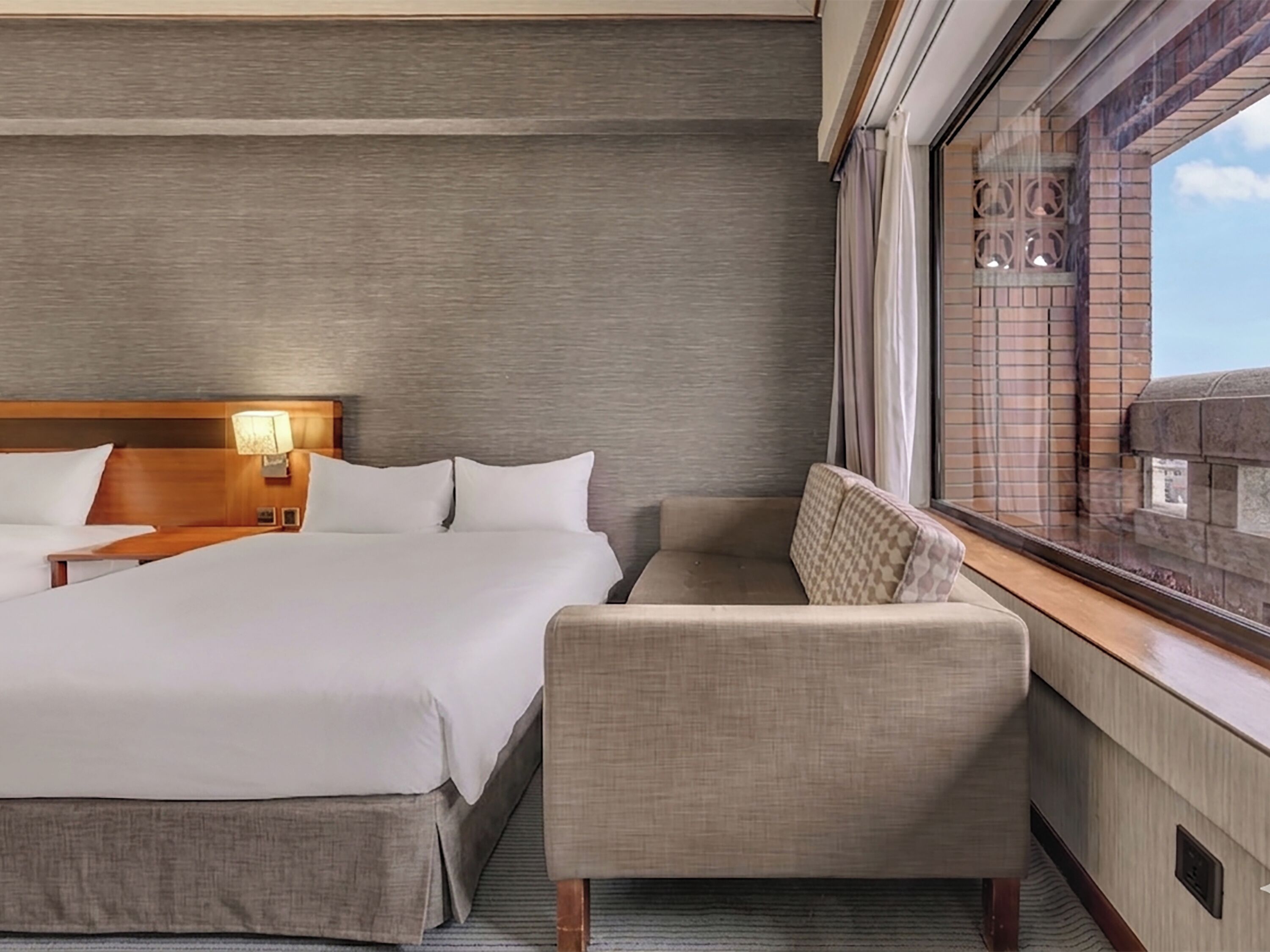 A hotel room with a bed, a sofa, a nightstand, a lamp, and a window with a view of the outside.