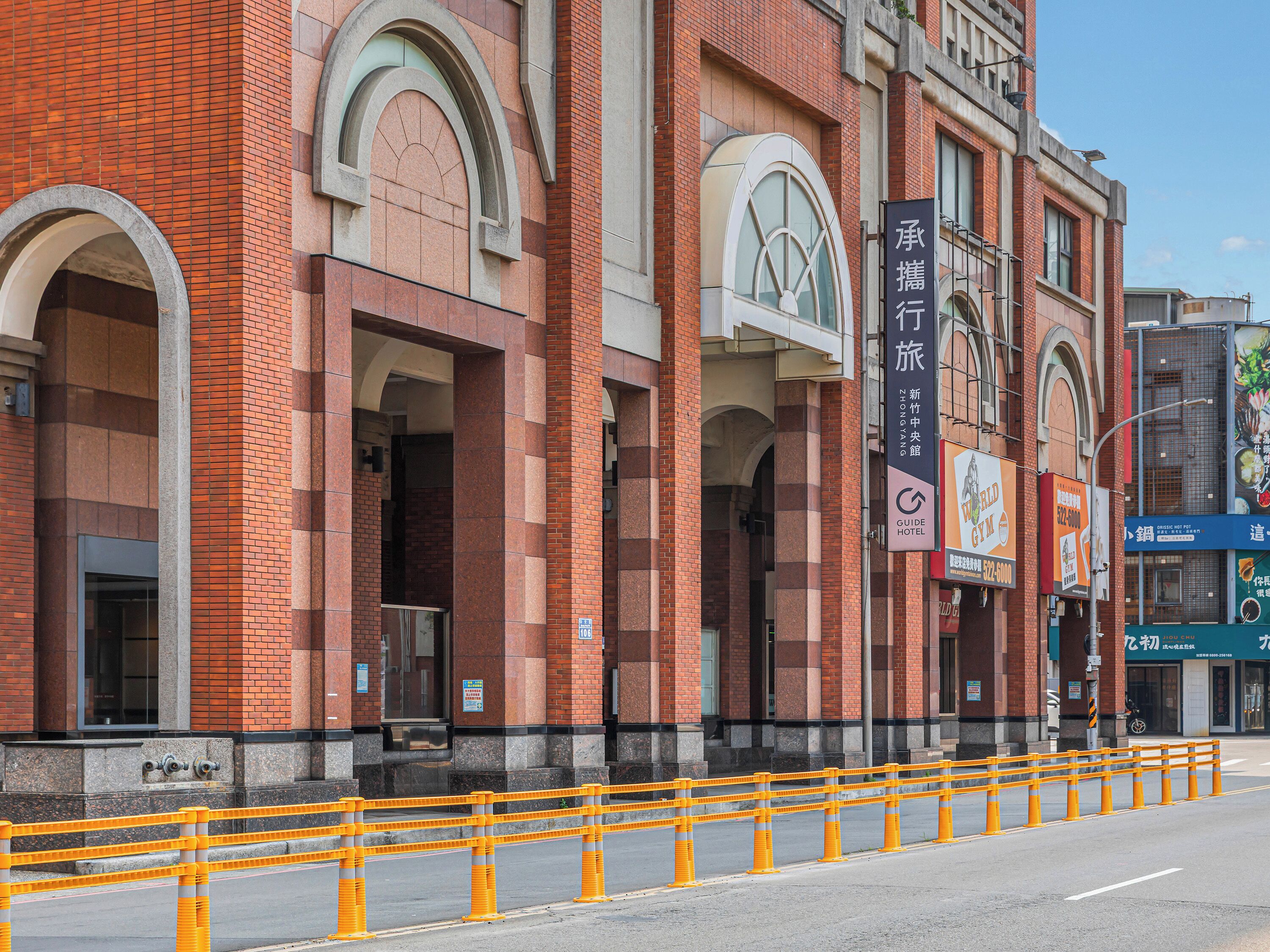A brick building with large arched windows and a yellow safety barrier in front.