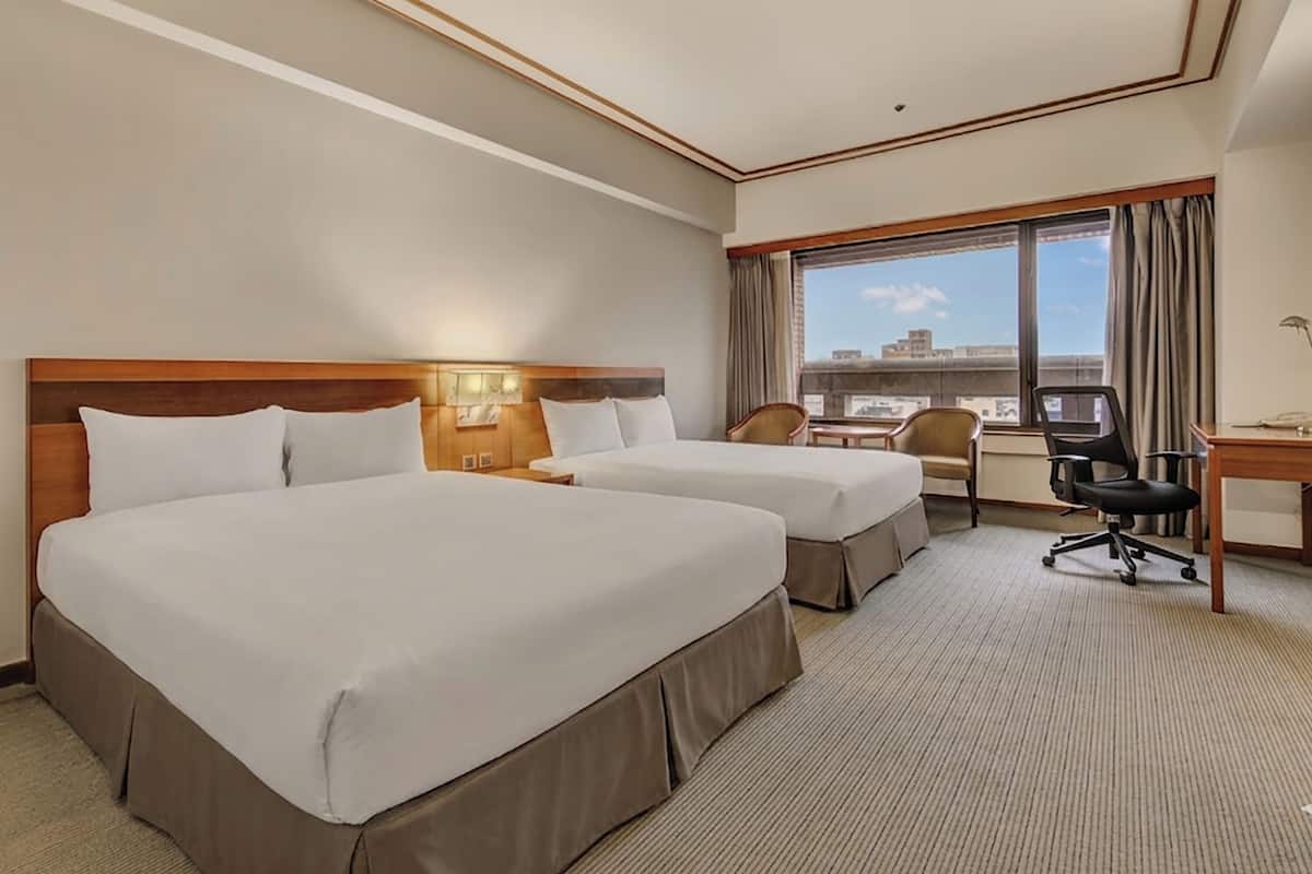 A hotel room with two beds, a desk, a chair, and a large window offering a city view.