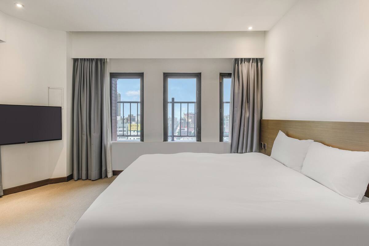 A hotel room with a large bed, a flat-screen TV mounted on the wall, and a city view through three windows.