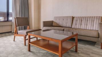 A hotel room with a sofa, a wooden coffee table, a chair, and a view of the city through a window.